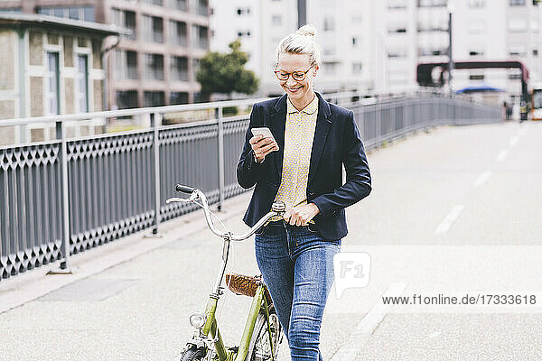 Blond female commuter wheeling bicycle while using mobile phone on bridge
