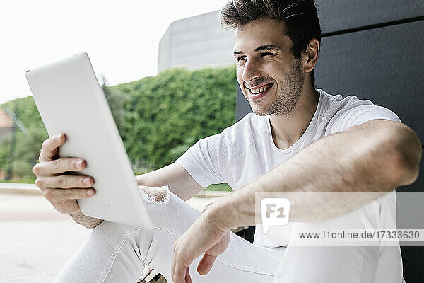 Junger Mann lächelt während eines Videoanrufs über ein digitales Tablet  während er vor einer Säule sitzt