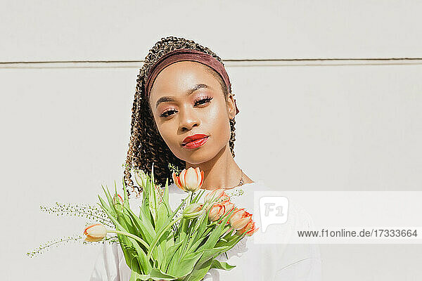 Beautiful woman with flowers standing in front of white wall on sunny day