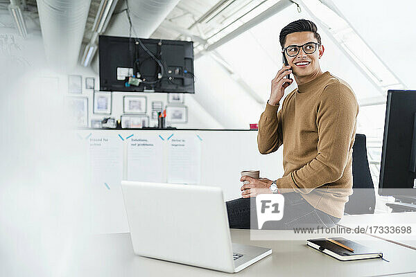 Smiling businessman talking on mobile phone while sitting by laptop in office