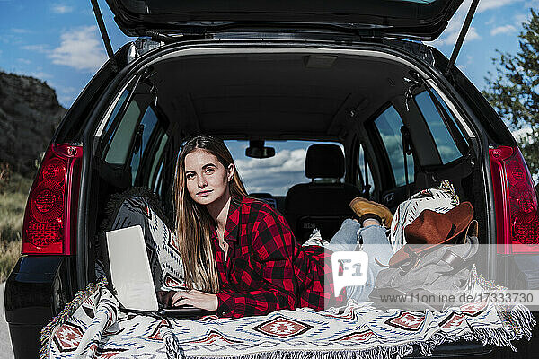 Female freelancer lying with laptop in car trunk on sunny day