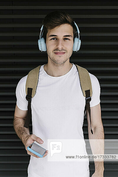 Junger Mann hält Handy und hört Musik über Kopfhörer vor einem Fensterladen