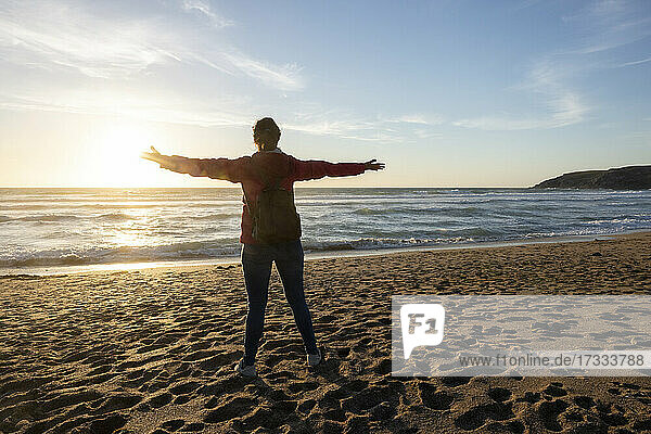 Frau steht mit ausgestreckten Armen am Strand bei Sonnenuntergang