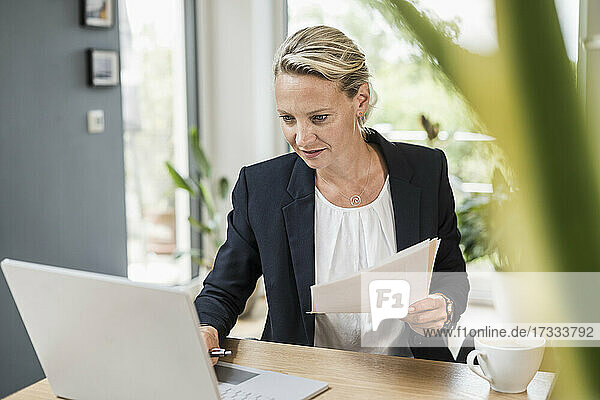 Geschäftsfrau hält ein Dokument  während sie einen Laptop im Büro benutzt