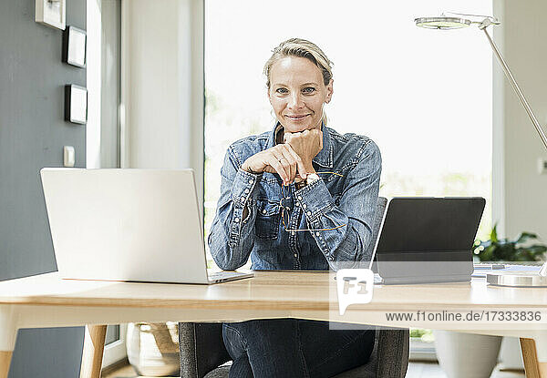 Smiling female freelancer with laptop and digital tablet sitting with hand on chin at home office
