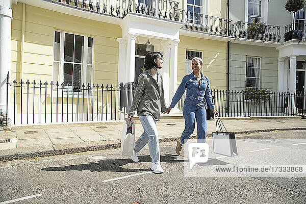 Lächelnde Freundinnen mit Einkaufstaschen beim Überqueren der Straße in der Stadt an einem sonnigen Tag
