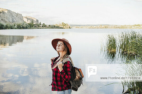 Young beautiful woman wearing hat standing with eyes closed at lakeshore