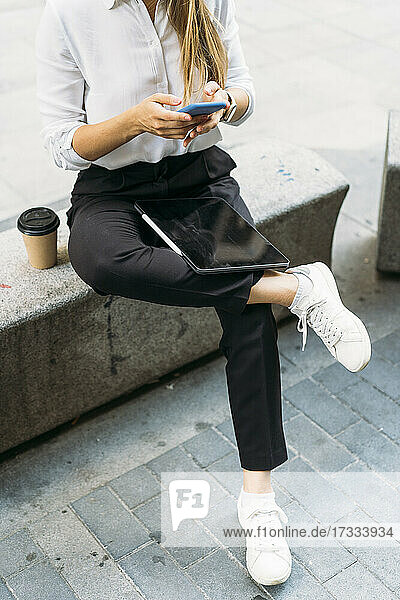 Geschäftsfrau  die auf einer Bank sitzend eine Textnachricht über ihr Smartphone verschickt