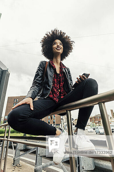 Lächelnde Frau mit Mobiltelefon auf dem Geländer sitzend