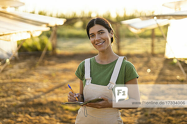 Lächelnde junge Bäuerin mit Notizblock auf einem Bauernhof an einem sonnigen Tag