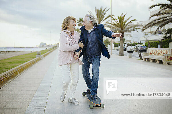 Fröhlicher reifer Mann  der mit dem Skateboard an einer Frau vorbeifährt  die sich auf dem Gehweg an den Händen hält