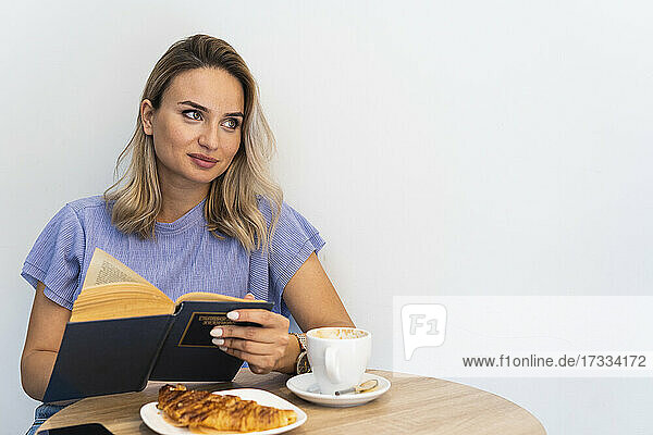 Junge Frau mit Buch  die wegschaut  während sie an einem Tisch vor einer Wand in einem Café sitzt