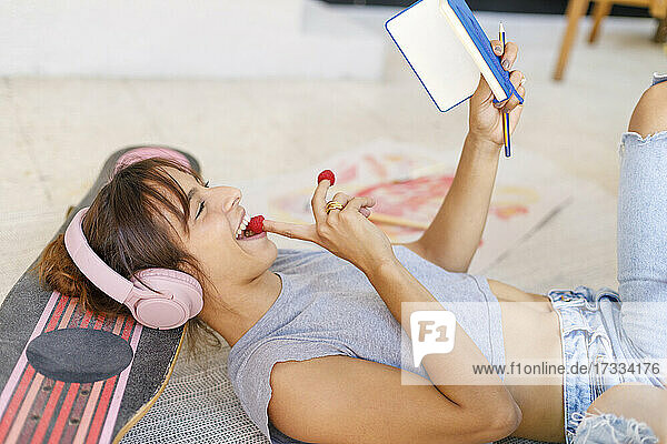 Happy female creative professional eating raspberry while reading notepad in studio