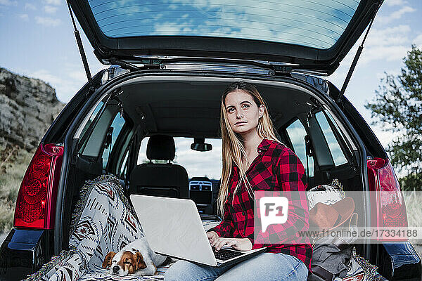 Junge blonde digitale Nomadin sitzt mit Laptop und Hund im Kofferraum