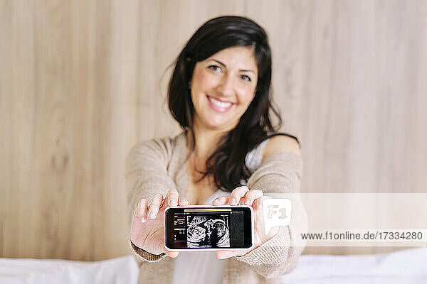 Lächelnde schwangere Frau zeigt Ultraschall auf dem Mobiltelefon