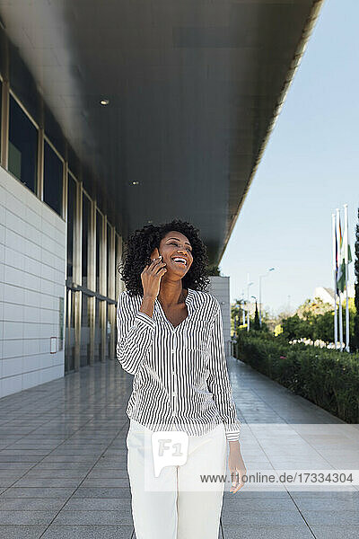 Junge Frau lacht  während sie mit einem Mobiltelefon spricht  das vor einem Bürogebäude steht