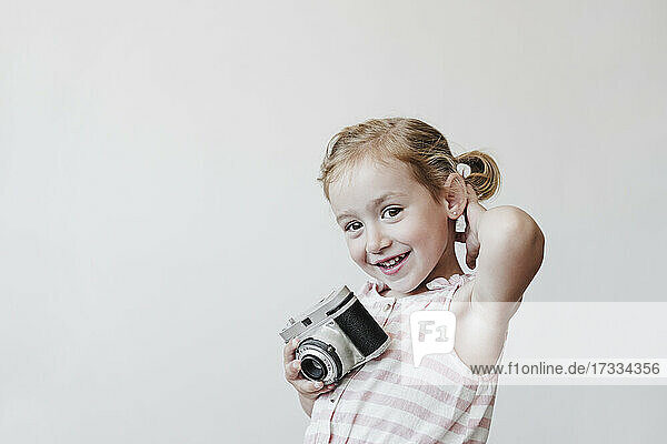 Lächelndes blondes Mädchen hält Spielzeugkamera vor weißem Hintergrund