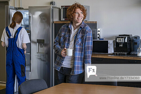 Smiling apprentice having coffee while female colleague searching in refrigerator at cafeteria