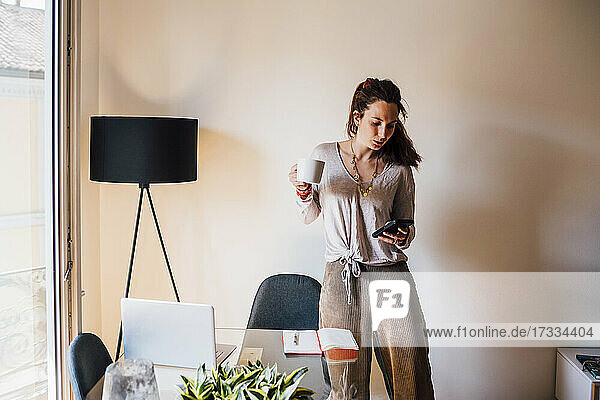 Woman holding coffee cup while using mobile phone at home