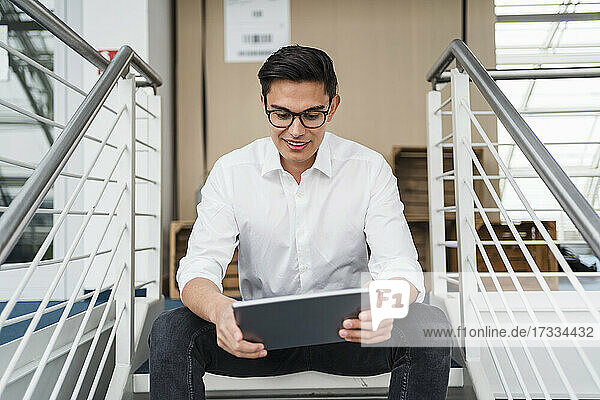 Lächelnder Geschäftsmann  der ein digitales Tablet benutzt  während er auf einer Treppe sitzt