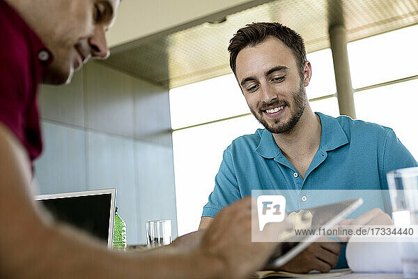 Mittlerer erwachsener Geschäftsmann  der über ein digitales Tablet mit einem lächelnden männlichen Kollegen im Büro diskutiert