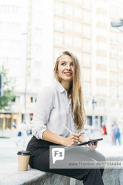 Lächelnde Geschäftsfrau mit digitalem Tablet und Einweg-Kaffeebecher auf einer Bank sitzend