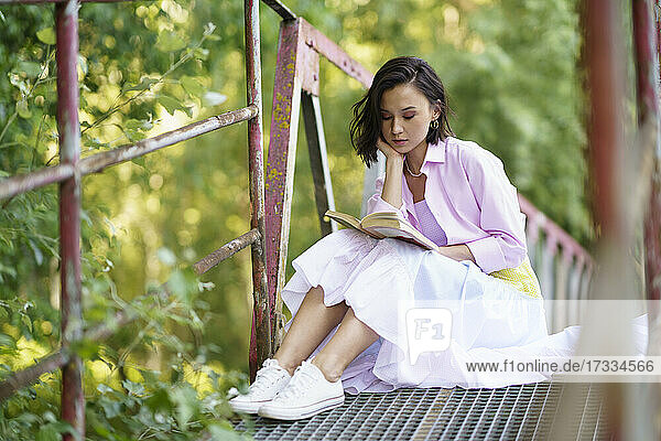 Junge Frau sitzt mit der Hand am Kinn und liest ein Buch auf einer Fußgängerbrücke auf dem Lande