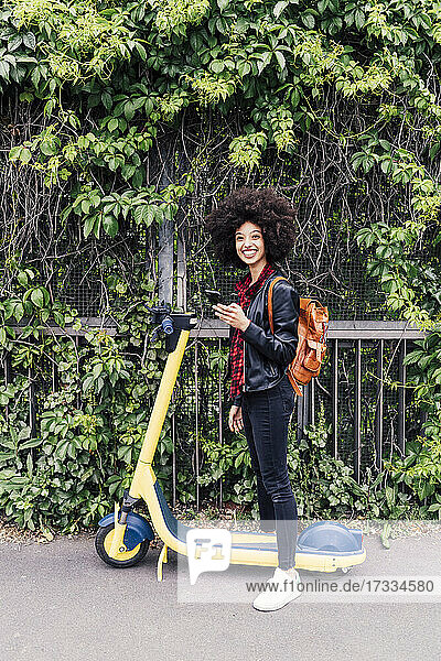 Smiling woman holding mobile phone while standing with electric push scooter on road