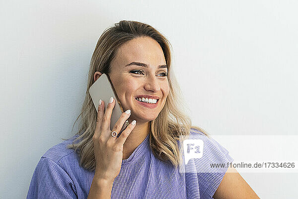 Lächelnde junge Frau  die wegschaut  während sie mit einem Handy vor einer Wand spricht