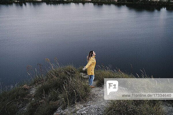 Junge Frau in gelbem Regenmantel steht mit ausgestreckten Armen am Seeufer