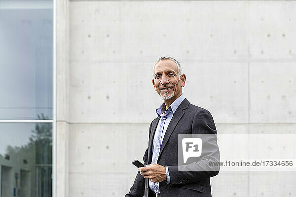 Smiling businessman holding smart phone near office building