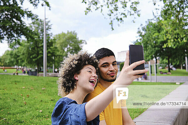 Glückliche junge Frau  die ein Selfie mit ihrem Freund über ihr Smartphone macht  während sie im Park sitzt