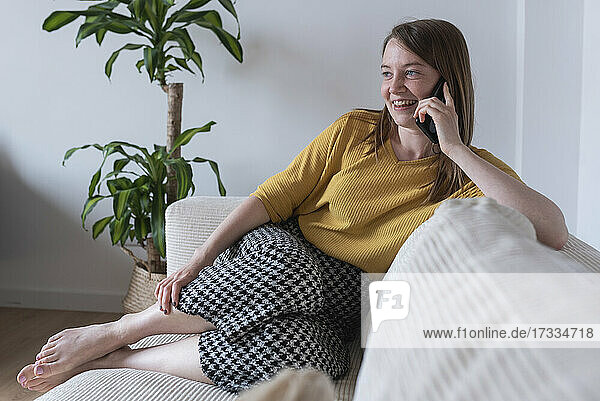 Lächelnde Frau auf dem Sofa sitzend  während sie zu Hause mit ihrem Smartphone telefoniert