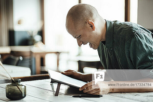 Glücklicher glatzköpfiger Geschäftsmann  der in einem Café sitzt und auf ein digitales Tablet schaut