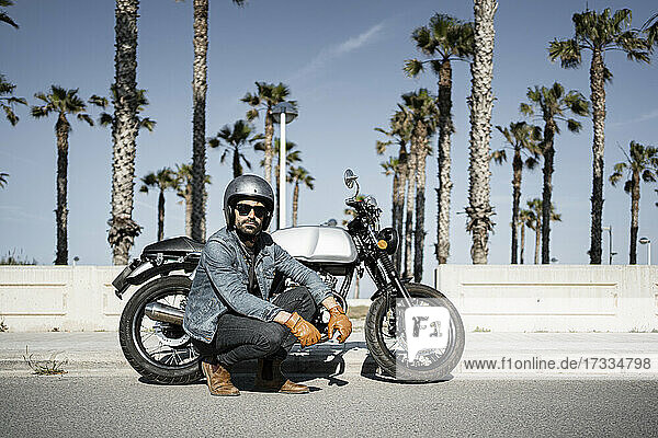 Handsome man squatting by motorcycle on sunny day