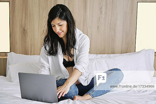 Schwangere Frau im mittleren Erwachsenenalter  die einen Laptop benutzt  während sie auf dem Bett im Schlafzimmer sitzt