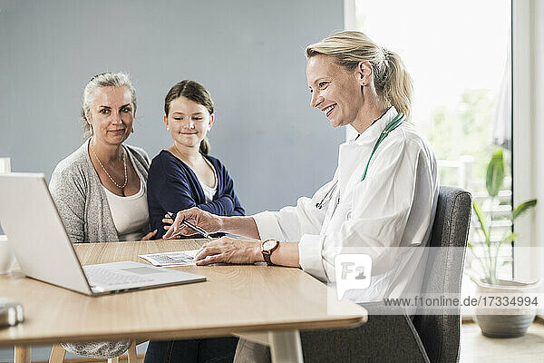 Lächelnder Kinderarzt  der auf einen Laptop schaut  während er mit einem Mädchen und einer Frau im Büro sitzt