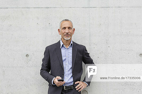 Businessman with hand on hip holding smart phone near wall