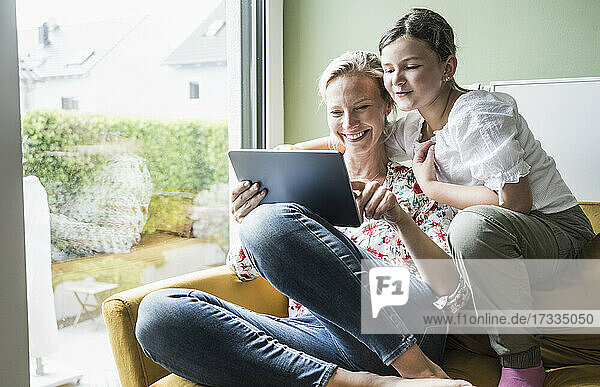Smiling mother and daughter using digital tablet while sitting on sofa