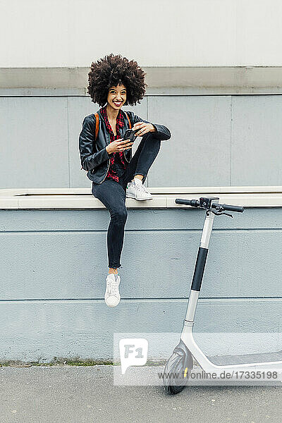 Smiling woman holding mobile phone while sitting on retaining wall by electric push scooter