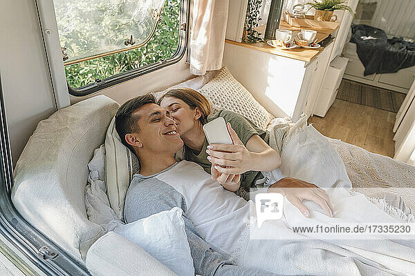 Woman kissing boyfriend while taking selfie on bed in motor home