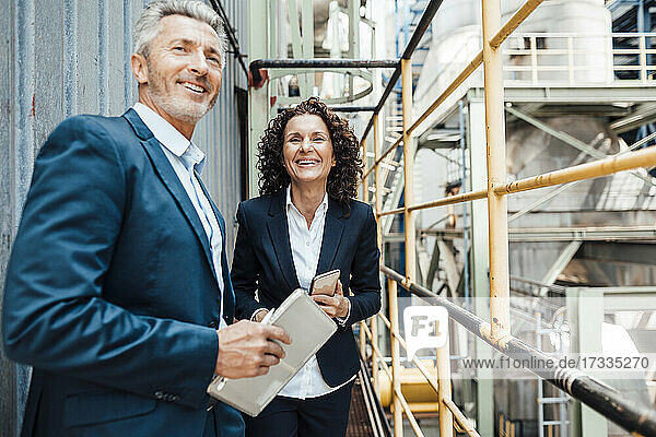 Lächelnde männliche Fachkraft  die ein digitales Tablet hält  während sie neben einem Kollegen in der Industrie steht
