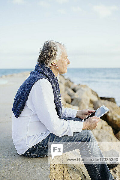 Mature man listening music through in-ear headphones while sitting at seaside