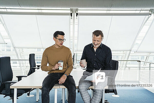 Male colleagues with coffee cup working while sitting on desk