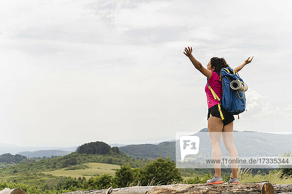 Carefree female backpacker with arms outstretched standing on log in front of sky