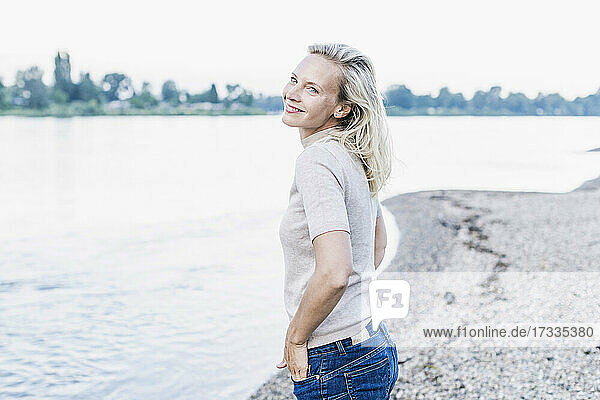 Lächelnde Frau mit Hand in der Tasche am Flussufer stehend