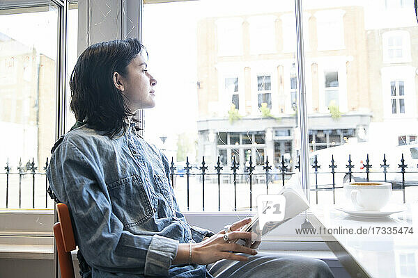 Junge Frau sitzt mit einem Buch im Café und schaut durch das Fenster