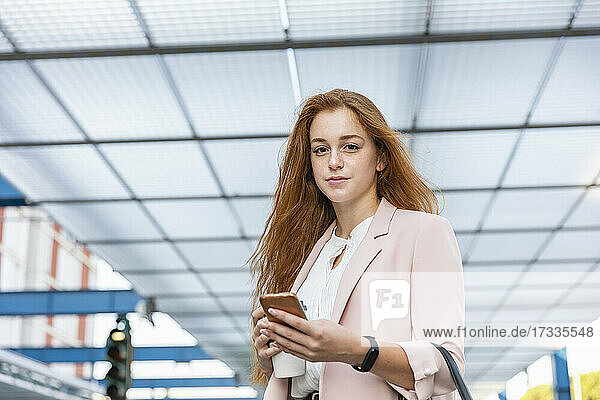 Rothaarige Geschäftsfrau mit Mobiltelefon und Einweg-Kaffeebecher im Schatten auf dem Bahnsteig