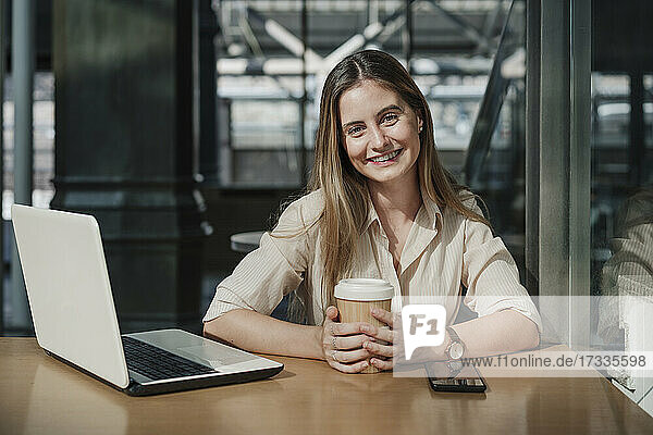 Glückliche junge Geschäftsfrau mit Einweg-Kaffeebecher in einem Café sitzend