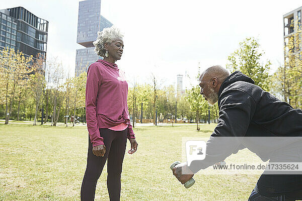 Ältere Frau schaut auf Mann  der sich beim Training mit Hanteln im Park bückt